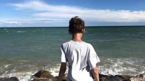 Boy on stone beach looking at sea water and drawing by stick Stock Footage 102333451