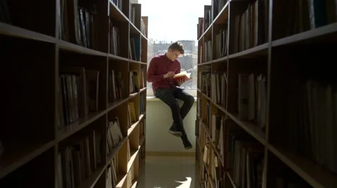 Boy -  student reading a book on a window sill at the library Stock Footage
