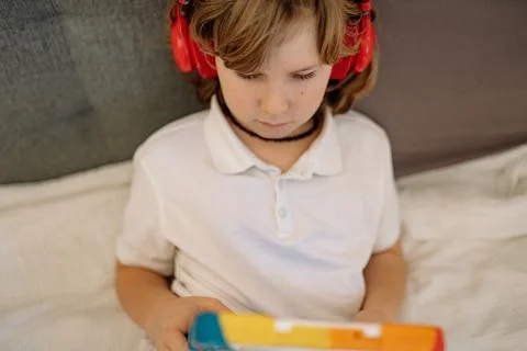 Boy studies on tablet in bedroom while seated on bed and wearing headphones Stock-Fotos
