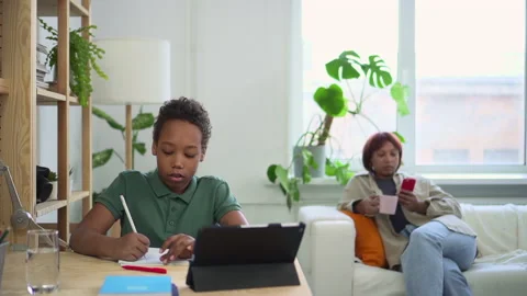 Boy studying in front of tablet and writing, mother using phone and sitting on Stock Footage 162731978