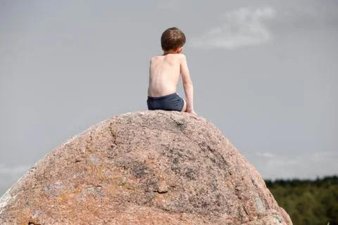 Boy sunbathes while sitting on a large, sloping, granite boulder Stock Photos