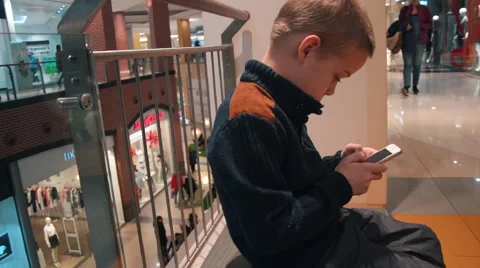 Boy in a Sweater is Playing in the Smartphone While His Parents Are Shopping at Stock Footage 61069353