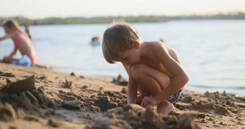 Boy in swim trunks digging sand with blue plastic shovel on beach near water Vidéo 306527505