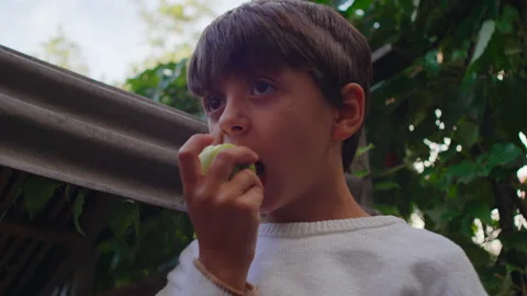 Boy taking bite of fresh apple in leafy outdoor setting creating natural moment Stock Footage 323702143