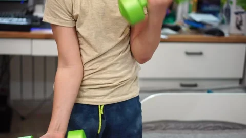 Boy ten years old pumping biceps using two green dumbbells standing in room. clo 库存影片 307488879