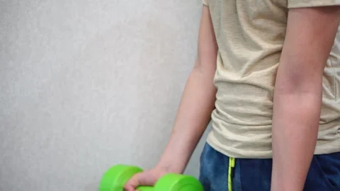 Boy ten years old pumping biceps using two green dumbbells standing in room. clo Video stock 307488914