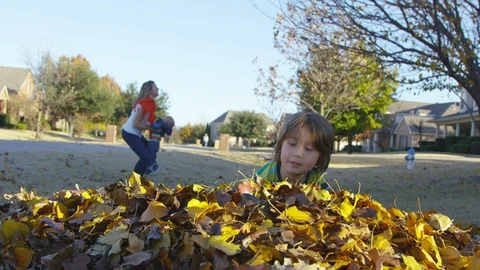 Boy throws leaves into the camera while mother plays with toddler in background Видео 94007969