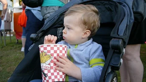 Boy toddler eats French fries from bucket on a festival sitting in stroller Stock Footage