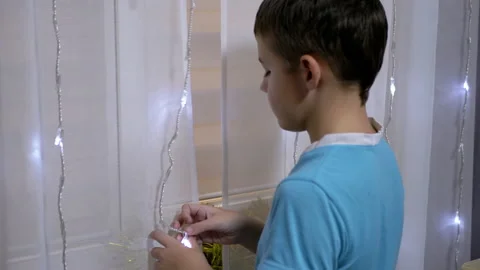 Boy Unwinds Tangled New Year's Garland on Curtain, Standing Near Window. Stock Footage 145244703
