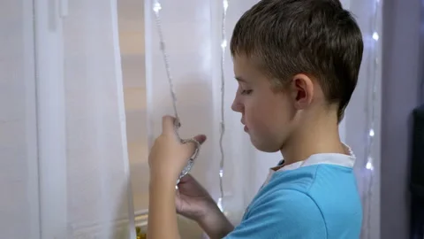 Boy Unwinds Tangled New Year's Garland on Curtain, Standing Near Window. Stock Footage 145605980