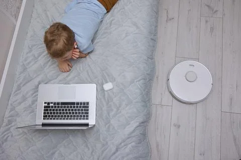 Boy uses a laptop on the bed while the robot vacuum cleaner does the cleaning Stock Photos