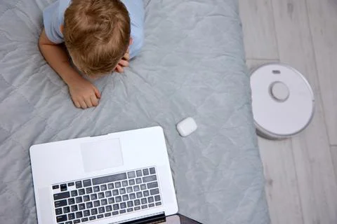 Boy uses a laptop on the bed while the robot vacuum cleaner does the cleaning Stock Photos