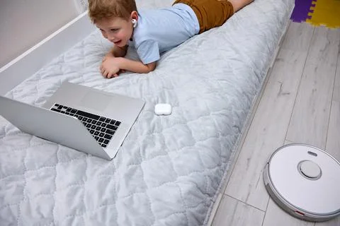Boy uses a laptop on the bed while the robot vacuum cleaner does the cleaning Stock Photos