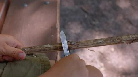 The boy uses a penknife to remove the bark from a stick. Close up. Foto stock
