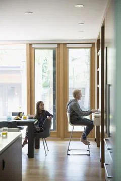 Boy using computer at desk and girl eating breakfast in kitchen Stock Photos