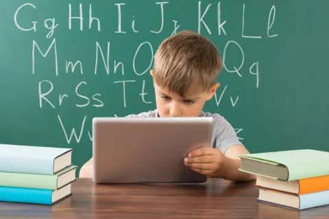 Boy Using Digital Tablet With Stack Of Books On Desk In Classroom 写真素材