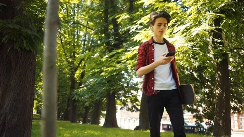 Boy using mobile phone listens to music in headphones walk keeps skate in park Stock Footage 92049111