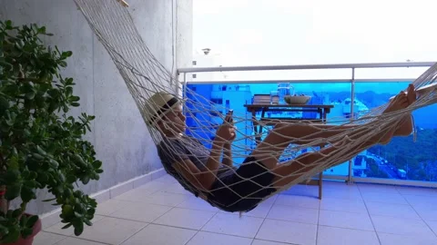 Boy using mobile phone lying in the hammock hanging on balcony of apartment Stock Footage 133443292