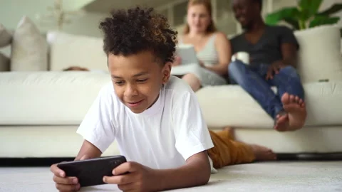 Boy using smartphone and lying on floor, parents sitting on couch in home room Stock Footage 152324510