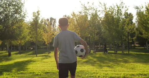 Boy walks across green grass in sunny park, holding soccer ball in one hand Stock Footage 346690495