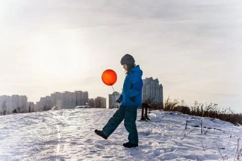 A boy in a winter blue jacket and hat stands with a red balloon on a stick Stock Photos