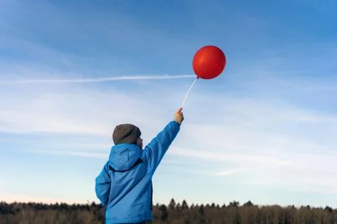 A boy in a winter blue jacket and hat stands with a red balloon on a stick Stock-Fotos
