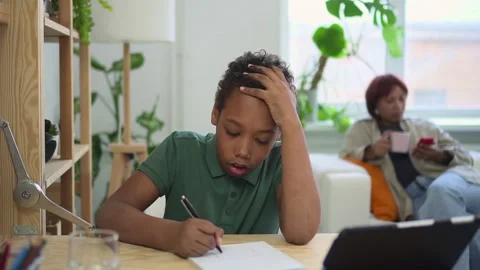 Boy writing in notebook and studying, sitting at table with tablet at home, mom Stock Footage 162731907