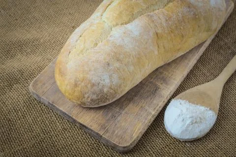 Bread and spoon filled with flour on bread board on burlap background Foto stock
