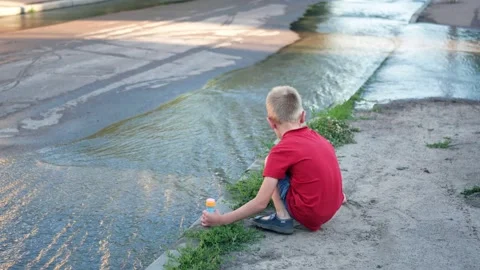 Break of the water pipe. Water flows down a city street and a child boy play Stock Footage 144078242