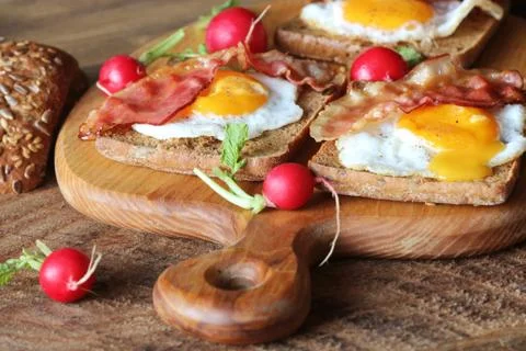 Breakfast of crispy bacon, fried eggs and bread. Sandwiches on cutting board. Stock Photos