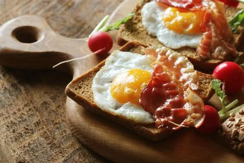 Breakfast of crispy bacon, fried eggs and bread. Sandwiches on cutting board. Stock Photos