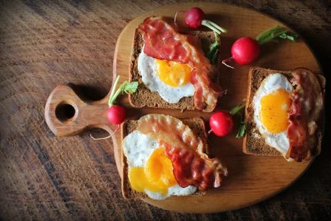 Breakfast of crispy bacon, fried eggs and bread. Sandwiches on cutting board. Stock Photos