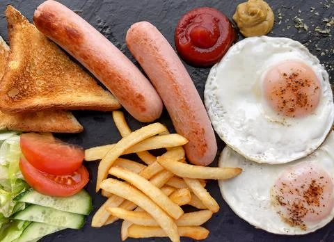 Breakfast on dark board with salad, eggs, sausages, french fries and toasts.. Stock Photos