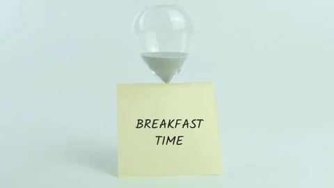 Breakfast time written on yellow sticky note placed on hourglass on white Stock Footage 313463969