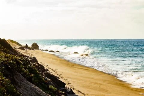 Breaking wave on sandy beach, man walking leaving footprints into distance Stock Photos
