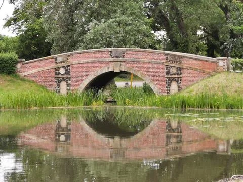 The brick bridge is reflected in the pond located in the green park 스톡 사진