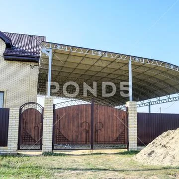 Brick house with a fence and gates. A large awning with a steel frame. View o Stock Photos