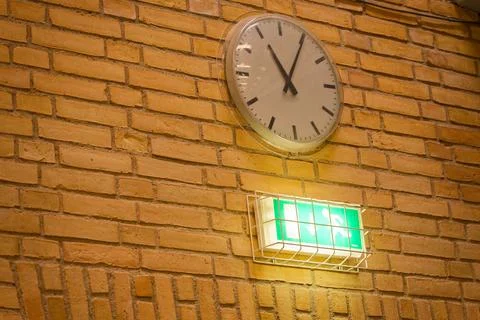 Brick wall clock under cover with caged exit light below. Stock Photos