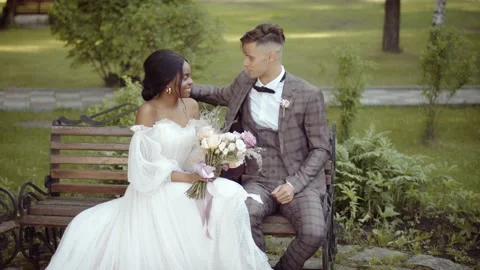 The bride and groom of different races sit side by side on the bench, chatting Vidéo 156256997