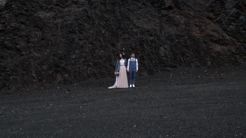Bride and groom in front of contrast dark rock. Newlyweds holding hands and turn Vidéo 101977353