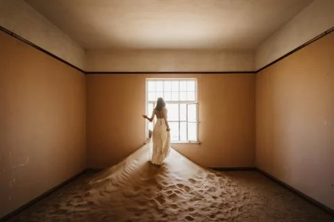 Bride looking outside windows of an abandoned desert villa filled with sand Foto stock
