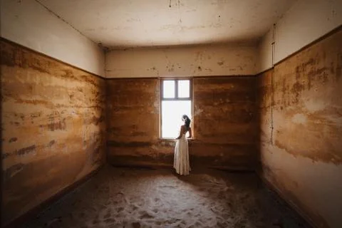 Bride looking outside windows of an abandoned desert villa filled with sand Foto stock