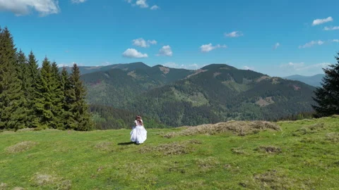 Bride sits alone in a vast alpine meadow beneath distant mountain peaks, Wide Stock Footage 346802397