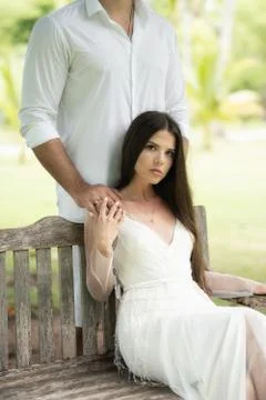 The bride sits on a bench, while the groom stands behind and holds her by the Stock Photos