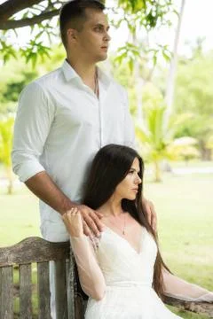 The bride sits on a bench, while the groom stands behind and holds her by the Stock Photos