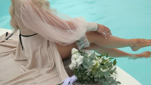 The bride sits in the pool, looking at her wedding bouquet and stroking her leg Stock Footage 235855666