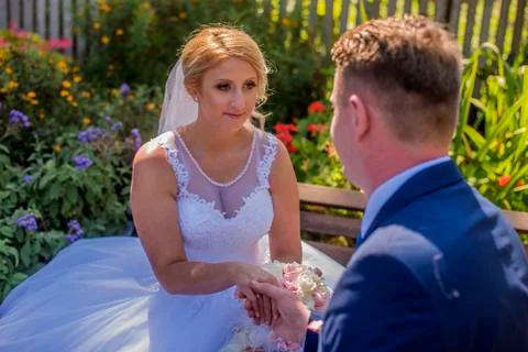 Bride sitting on garden bench while man is holding her hand. Stock Photos