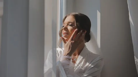 Bride Smiling by Sunlit Window in White Robe, A bride in a white robe smiles as Stock Footage 290778498