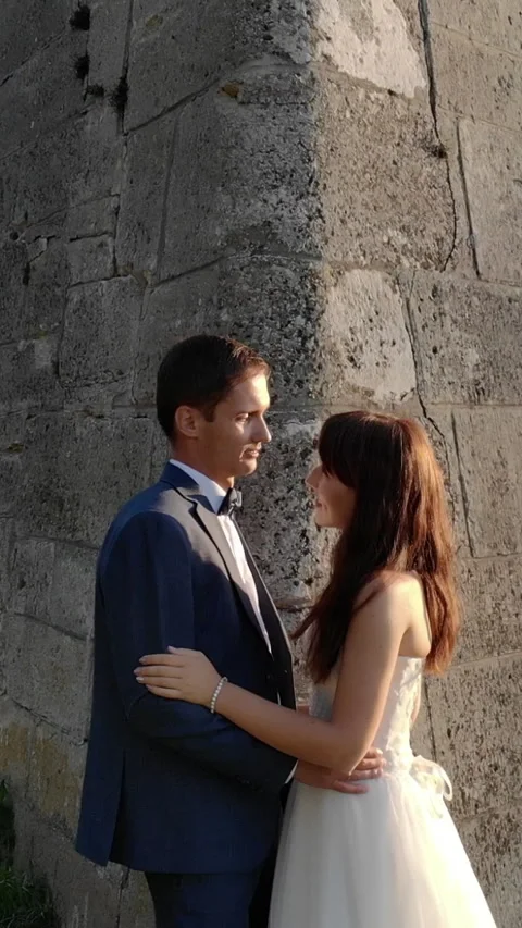 Bride stands by a stone wall, watching a couple kiss at sunset. Soft golden Stock Footage 313753857