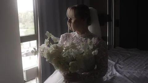 Bride in wedding dress sits on bed with bouquet by window Stock Footage 265583835